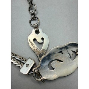 May include: Close-up of a silver-toned necklace with a heart-shaped pendant and a decorative, openwork design. The word "Brighton" is visible on the heart pendant. The chain has a woven design.