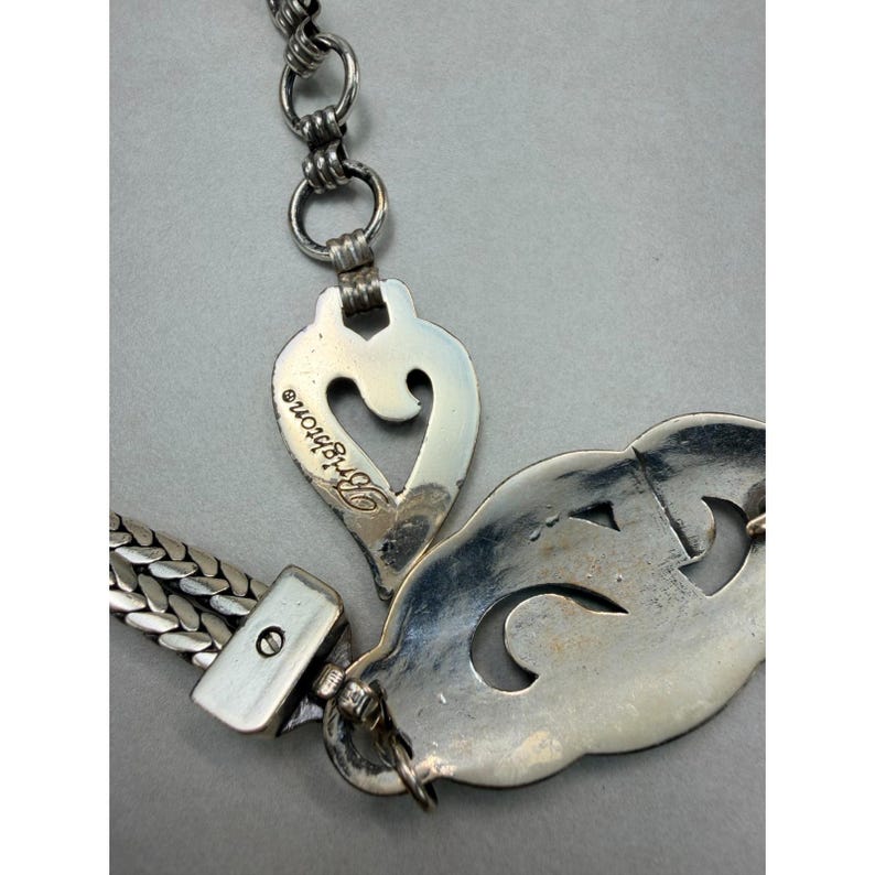 May include: Close-up of a silver-toned necklace with a heart-shaped pendant and a decorative, openwork design. The word "Brighton" is visible on the heart pendant. The chain has a woven design.