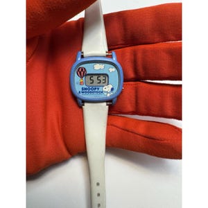May include: A vintage Snoopy & Woodstock wristwatch with a white band and a blue face. The watch face features a hot air balloon, clouds, and the time 5:53. The words "Snoopy & Woodstock" are printed on the watch face.