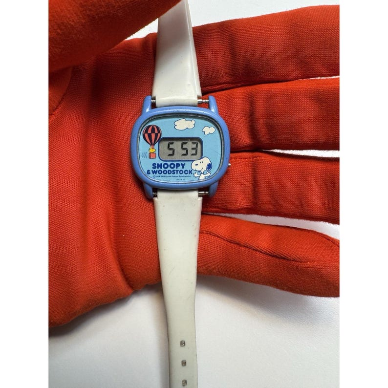 May include: A vintage Snoopy & Woodstock wristwatch with a white band and a blue face. The watch face features a hot air balloon, clouds, and the time 5:53. The words "Snoopy & Woodstock" are printed on the watch face.
