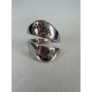May include: A silver-toned bypass ring featuring a polished finish. The upper portion of the ring displays an engraved mountain range design, while the lower portion features an engraved pattern of three evergreen trees on the metal surface.