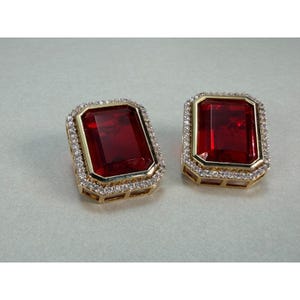 May include: A pair of rectangular earrings featuring large, deep red faceted stones set in gold-toned metal frames. Each central red stone is surrounded by a border of small, clear, sparkling white stones that line the outer edge of the setting.