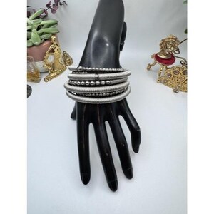 May include: A collection of silver-toned bracelets displayed on a black hand-shaped jewelry holder. The bracelets include multiple coiled bands and beaded strands. The jewelry holder is set against a white background with decorative items.