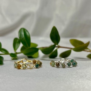 May include: Two wire-wrapped rings featuring green and brown agate stones. The ring on the left has a gold wire, and the ring on the right has a silver wire.