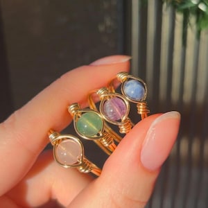 Peut inclure: Quatre bagues en fil de fer doré avec des perles de pierres précieuses de différentes couleurs. Les bagues sont empilées les unes sur les autres. Les perles sont rose clair, vertes, violettes et bleues.