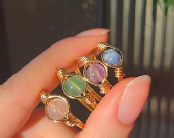 Pode incluir: Quatro anéis de arame dourado envoltos com contas de pedras preciosas de diferentes cores. Os anéis estão empilhados uns sobre os outros. As contas são rosa claro, verde, roxo e azul.