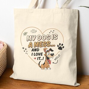 Puede incluir: Bolso tote de color crema con un gráfico en forma de corazón que dice "MY DOG IS A MESS... AND I LOVE IT..!" con una ilustración de perro de dibujos animados y el nombre "Milo". El bolso tiene dos asas y está hecho de tela resistente.