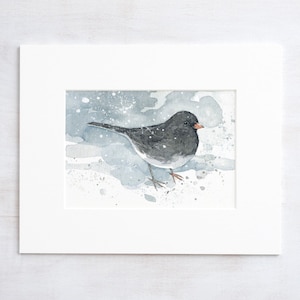 May include: Watercolor painting of a small bird with gray and white feathers, perched on a snowy surface. The bird has a small orange beak and is set against a backdrop of soft blue and gray watercolor washes, framed in a white mat.