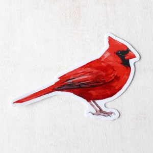 May include: A red cardinal bird sticker with a white background. The bird is facing right and has a black eye and beak.