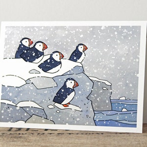 May include: A watercolor painting of five puffins perched on a snowy cliff overlooking a blue ocean. The puffins are black and white with orange beaks. The painting is set against a backdrop of a snowy day with falling snow.