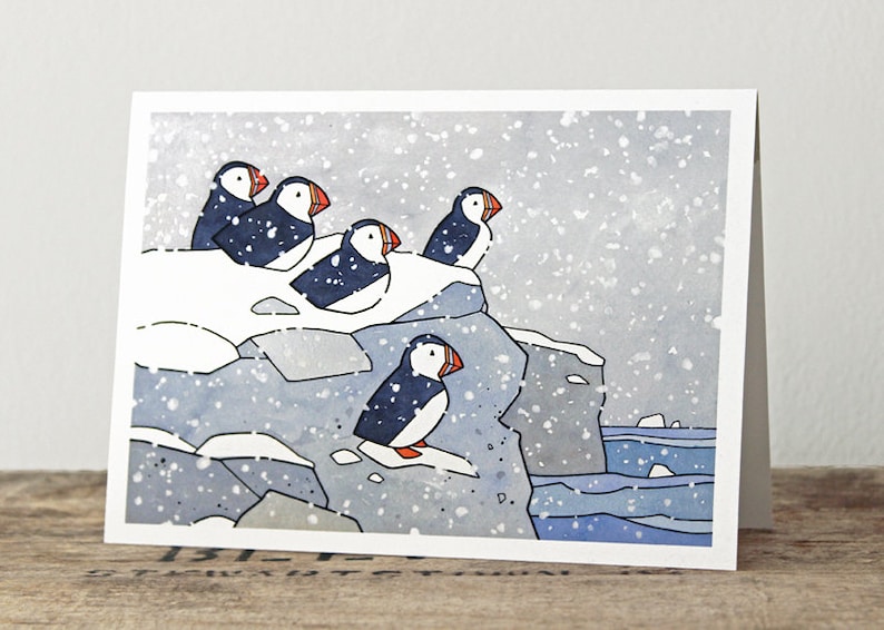 May include: A watercolor painting of five puffins perched on a snowy cliff overlooking a blue ocean. The puffins are black and white with orange beaks. The painting is set against a backdrop of a snowy day with falling snow.