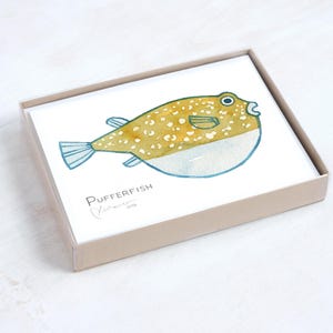 May include: A set of cards featuring a watercolor illustration of a pufferfish. The fish is yellow with white spots and blue accents, and is contained in a light brown box. The text "Pufferfish" and an artist's signature are visible.
