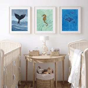 May include: Three framed watercolor paintings of sea creatures, including a whale tail, a seahorse, and a group of stingrays. The paintings are hung on a white wall above a wooden table with a lamp and baskets.