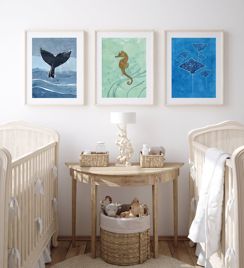 May include: Three framed watercolor paintings of sea creatures, including a whale tail, a seahorse, and a group of stingrays. The paintings are hung on a white wall above a wooden table with a lamp and baskets.