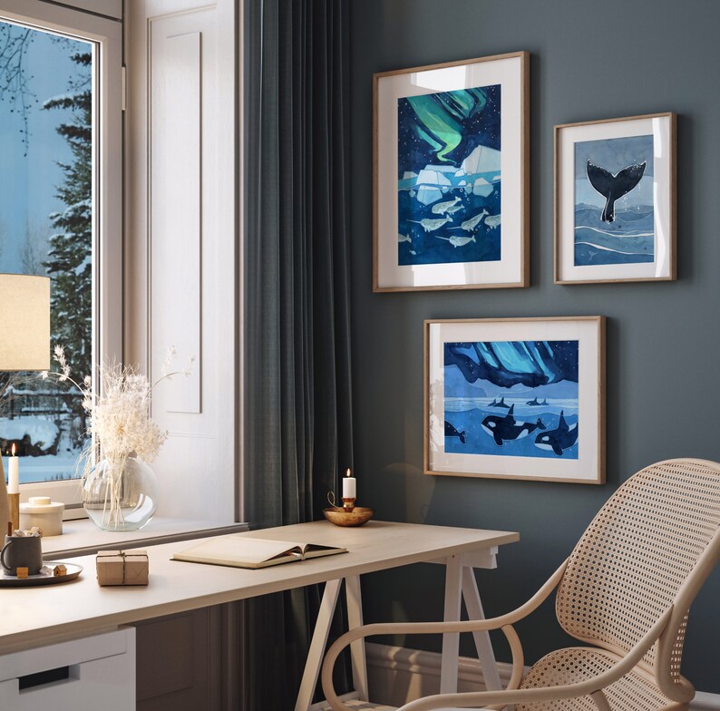 May include: Three framed illustrations of whales and the aurora borealis in a home office setting. The illustrations are in shades of blue and green, with a white background. The illustrations are hanging on a dark blue wall, above a white desk with a wooden chair.