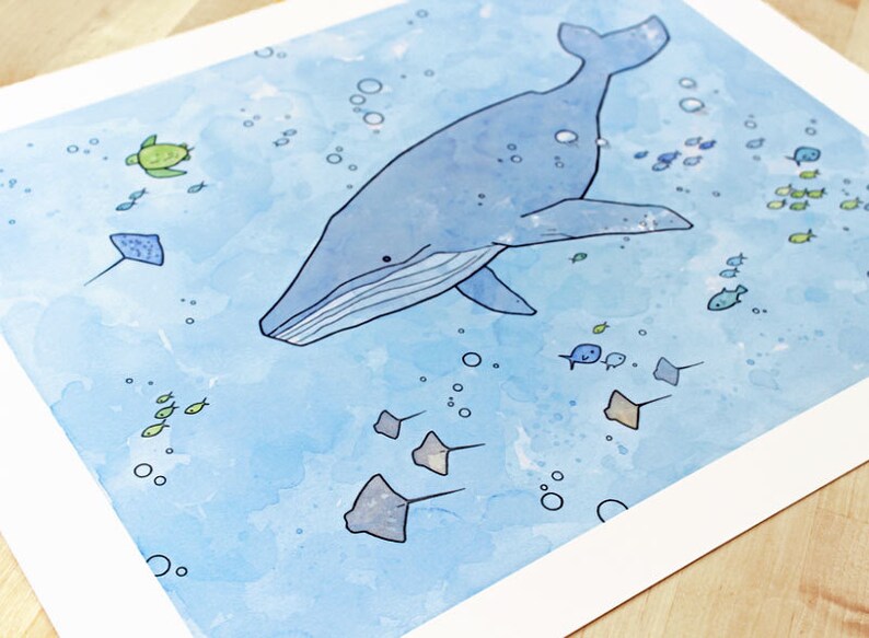 May include: A watercolor painting of a blue whale swimming in the ocean. The whale is surrounded by small fish, a sea turtle, and stingrays. The background is a light blue color with white bubbles.