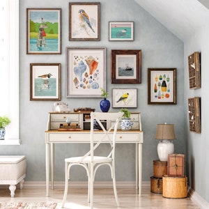 May include: A white desk with a wooden top and a white chair with a cross-back design. The desk has a variety of framed artwork on the wall behind it, including images of birds, seashells, and buoys. The artwork is in a coastal theme.
