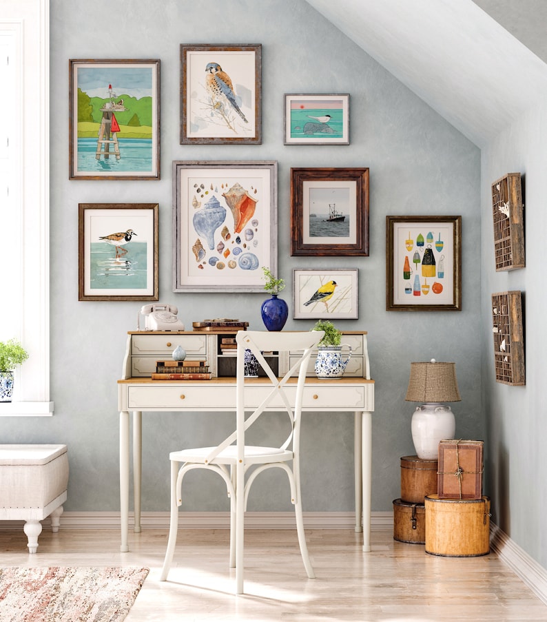 May include: A white desk with a wooden top and a white chair with a cross-back design. The desk has a variety of framed artwork on the wall behind it, including images of birds, seashells, and buoys. The artwork is in a coastal theme.