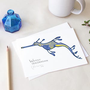 May include: A white card featuring a blue and yellow weedy seadragon illustration. The text "Weedy Seadragon" is below the illustration, with a signature and the year 2018. Two blue glass bottles, a pencil, and a mug are also visible.