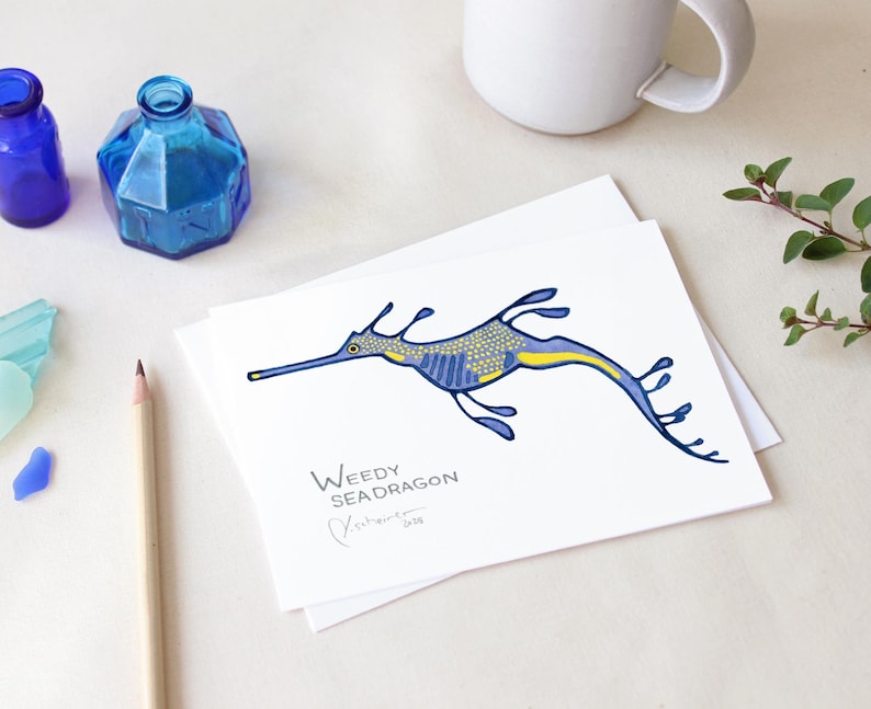 May include: A white card featuring a blue and yellow weedy seadragon illustration. The text "Weedy Seadragon" is below the illustration, with a signature and the year 2018. Two blue glass bottles, a pencil, and a mug are also visible.
