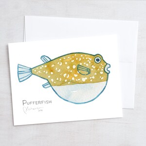 May include: Watercolor illustration of a pufferfish on a white card. The fish is yellow-brown with white spots and blue fins. The card is paired with a white envelope. The word "Pufferfish" is written below the fish.