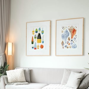 May include: Two framed watercolor illustrations, one depicting a collection of colorful buoys and the other featuring a variety of seashells. The illustrations are hung on a white wall above a gray couch with pillows.