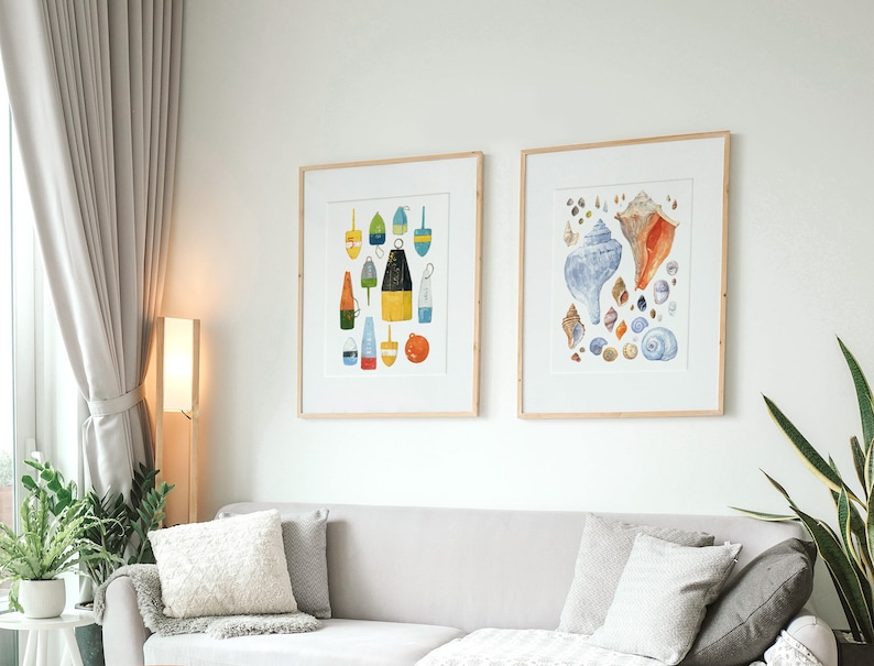 May include: Two framed watercolor illustrations, one depicting a collection of colorful buoys and the other featuring a variety of seashells. The illustrations are hung on a white wall above a gray couch with pillows.