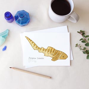 May include: A watercolor illustration of a zebra shark on a white card. The shark is tan with brown spots and stripes. The card is on a white surface with a pencil, a cup of coffee, and blue glass objects.