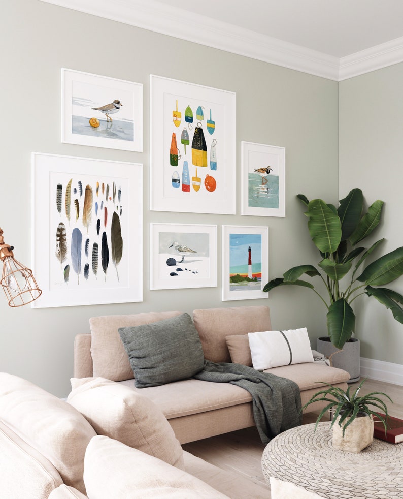 May include: A living room with a light beige sectional sofa, two throw pillows, a throw blanket, and a round woven coffee table. The wall behind the sofa is decorated with a gallery wall of framed prints featuring birds, feathers, and buoys. A potted plant is in the corner of the room.