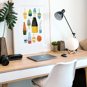 May include: A framed print of a colorful illustration of various buoy shapes and sizes. The buoys are painted in shades of blue, yellow, red, green, and white. The print is hanging on a white wall above a desk with a laptop, lamp, and other office supplies.
