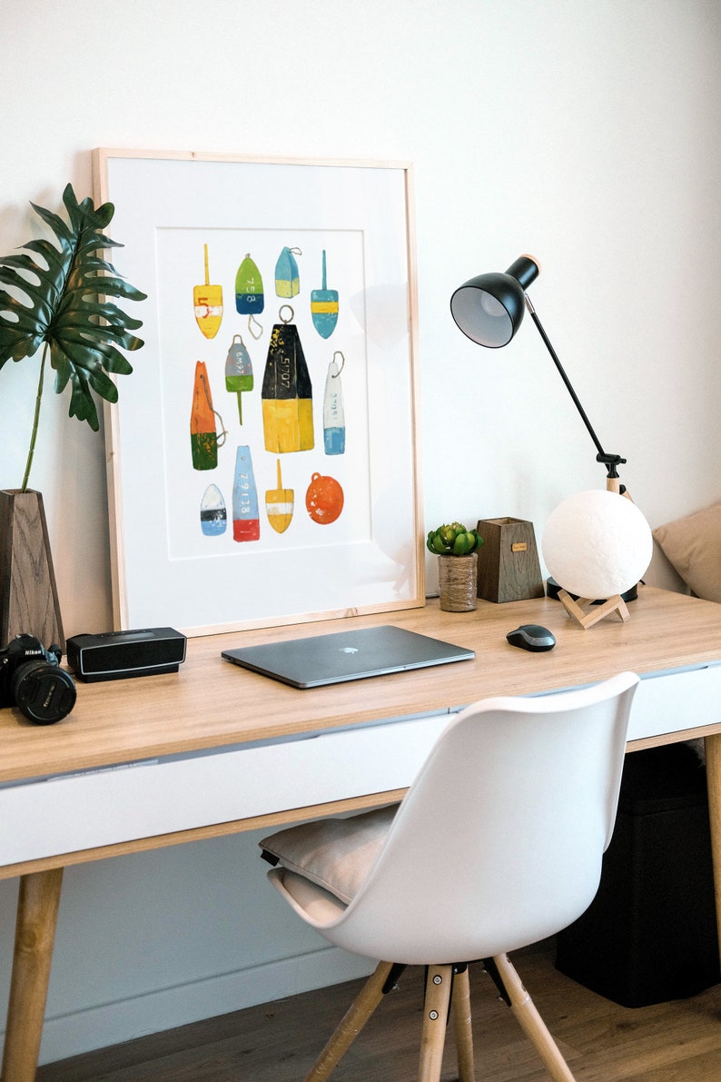 May include: A framed print of a colorful illustration of various buoy shapes and sizes. The buoys are painted in shades of blue, yellow, red, green, and white. The print is hanging on a white wall above a desk with a laptop, lamp, and other office supplies.