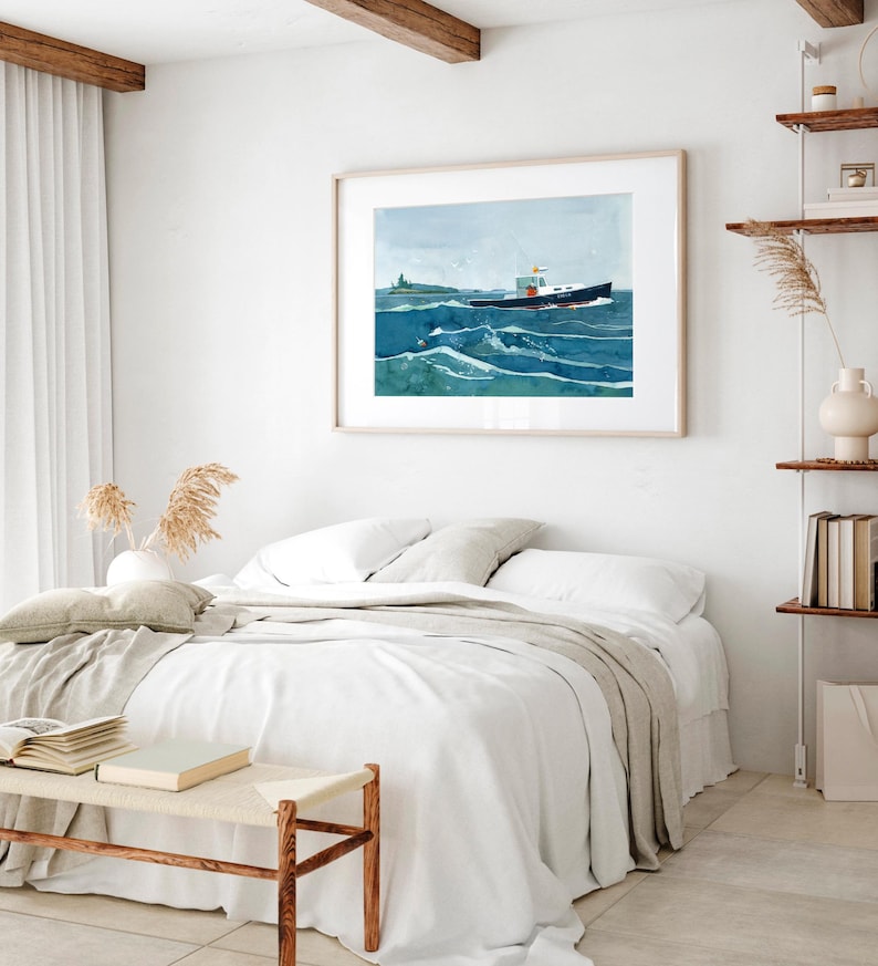 May include: A watercolor painting of a boat sailing on a blue ocean with white waves. The boat is dark blue with a white cabin and a red stripe. The painting is framed in a light wood frame and hangs on a white wall above a bed with white linens.