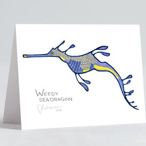 May include: A folded white greeting card featuring a blue and yellow illustration of a weedy seadragon. The seadragon has a long snout and flowing fins. The words "Weedy Seadragon" are printed below the illustration, along with an artist's signature.