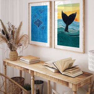 May include: Two framed watercolor paintings on a wooden console table. The left painting features a group of blue stingrays on a blue background. The right painting features a black whale tail emerging from turquoise water with a yellow sunset in the background.