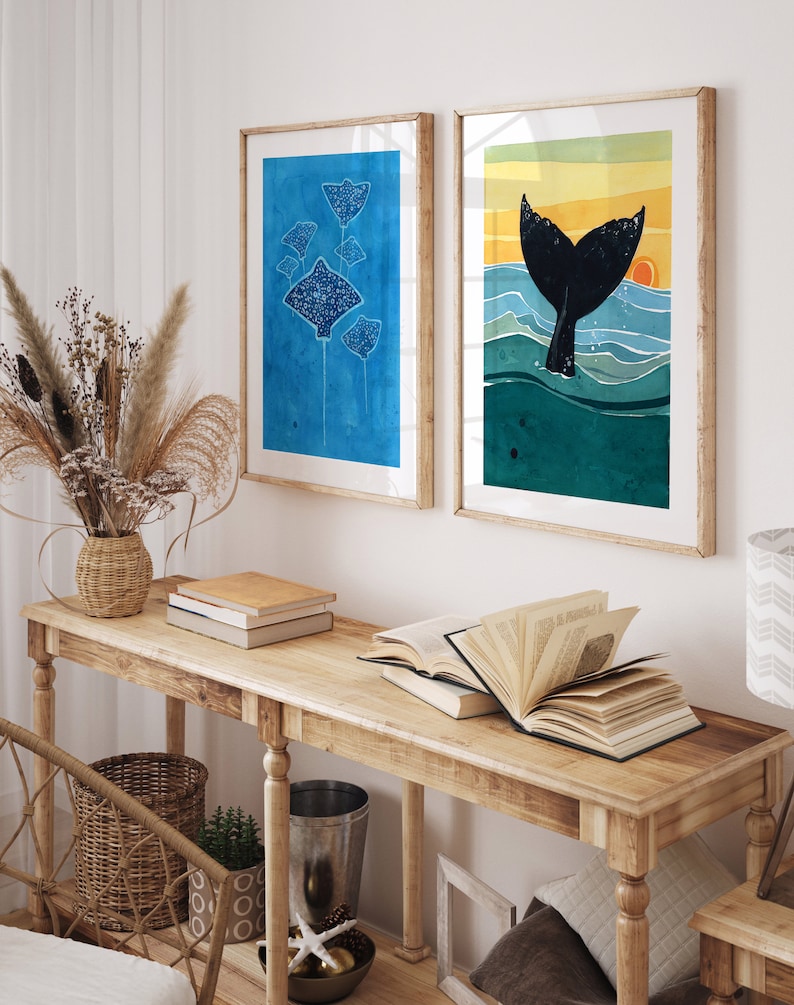 May include: Two framed watercolor paintings on a wooden console table. The left painting features a group of blue stingrays on a blue background. The right painting features a black whale tail emerging from turquoise water with a yellow sunset in the background.