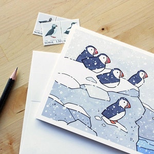 May include: A watercolor illustration of a group of puffins perched on a snowy cliff. The birds are facing different directions, with their beaks open and their wings folded. The background is a light blue color, with a few white dots representing snow.