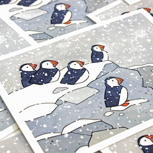 May include: A set of five illustrated greeting cards featuring puffins on a snowy cliffside. The cards are printed on white cardstock and have a watercolor-like style.
