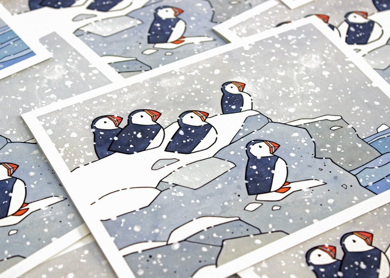 May include: A set of five illustrated greeting cards featuring puffins on a snowy cliffside. The cards are printed on white cardstock and have a watercolor-like style.