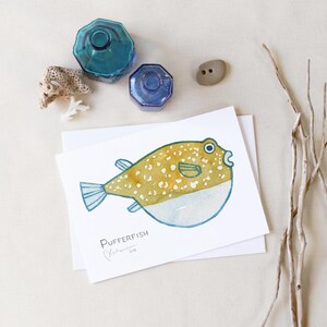 May include: Watercolor illustration of a pufferfish with yellow and white spots, blue fins, and a white belly. The artwork is on a white card with the word "Pufferfish" and artist's signature. Decorative blue glass bottles and a small stone are also in the image.
