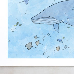 May include: A watercolor painting of a blue whale swimming in the ocean with a school of small fish and stingrays. The background is a light blue watercolor wash.