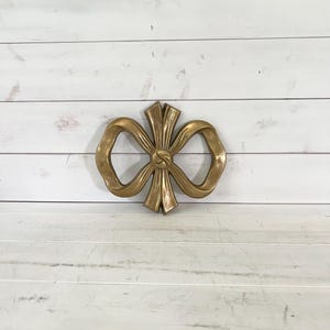 May include: A gold-coloured, bow-shaped wall decoration. The bow has a detailed knot in the centre and looped ends. The wall decor is made of a solid material and is mounted on a white wooden surface.