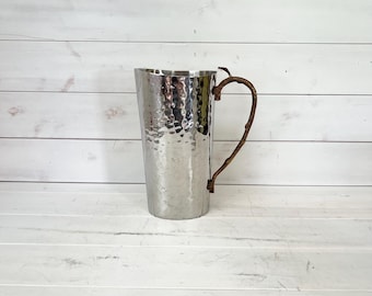 May include: A hammered metal pitcher with a natural twig handle. The silver-colored pitcher has a textured surface and a curved handle made from a brown twig. The pitcher is designed for serving beverages.