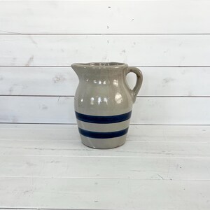 May include: A gray ceramic pitcher with a handle and spout, featuring two dark blue horizontal stripes around the body. The pitcher has a rustic, vintage aesthetic, suitable for kitchen or farmhouse decor.