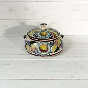 May include: A ceramic lidded bowl with a floral pattern. The bowl is cream-coloured with blue, yellow, and green flowers. It has two small handles and a knob on the lid. The rim is dark brown.