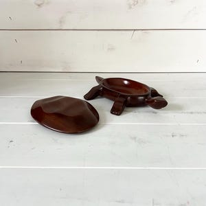 Puede incluir: Una caja de baratijas de madera con forma de tortuga y tapa. La caja y la tapa son de color marrón oscuro. La tortuga tiene un diseño detallado con patas y caparazón visibles. La tapa tiene forma ovalada y lisa.