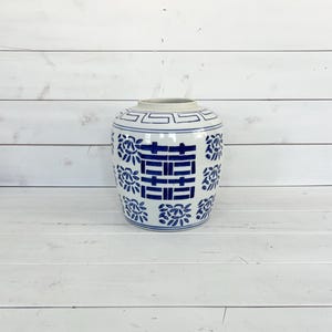 May include: A blue and white ceramic ginger jar with a geometric and floral pattern. The jar features a central blue double happiness symbol. The jar is cylindrical with a slightly flared rim and sits on a white wooden surface.