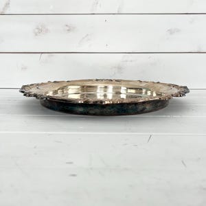 May include: A silver-colored, antique serving tray with an ornate, scalloped edge. The tray has a slightly raised center and a dark patina around the base, suggesting age and use. The background is a white, wooden surface.
