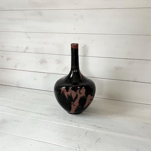 May include: A black ceramic vase with a brown glaze and a narrow neck. The vase has a rounded, bulbous body and is sitting on a white wooden surface.