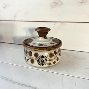 May include: A white ceramic jar with a brown lid and floral design. The jar has a brown and white floral pattern with a black centre.