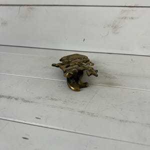 Może przedstawiać: Mała, mosiężna figurka przedstawiająca osobę grającą na instrumencie muzycznym. Figurka jest szczeg&oacute;łowa i ma błyszczące, metaliczne wykończenie. Figurka jest umieszczona na białej drewnianej powierzchni.